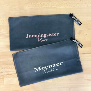 Pu&ograve; includere: Due asciugamani in microfibra grigi con testo bianco. L'asciugamano superiore dice "Jumpingsister Karo" in testo rosa. L'asciugamano inferiore dice "Meenzer Madche" in testo bianco.