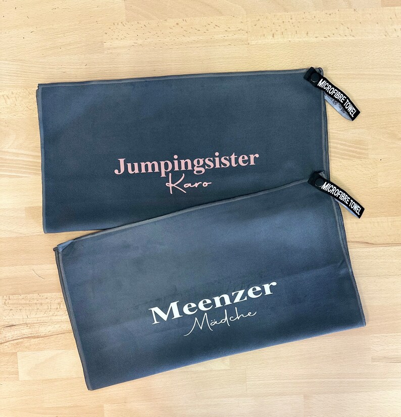 Pu&ograve; includere: Due asciugamani in microfibra grigi con testo bianco. L'asciugamano superiore dice "Jumpingsister Karo" in testo rosa. L'asciugamano inferiore dice "Meenzer Madche" in testo bianco.