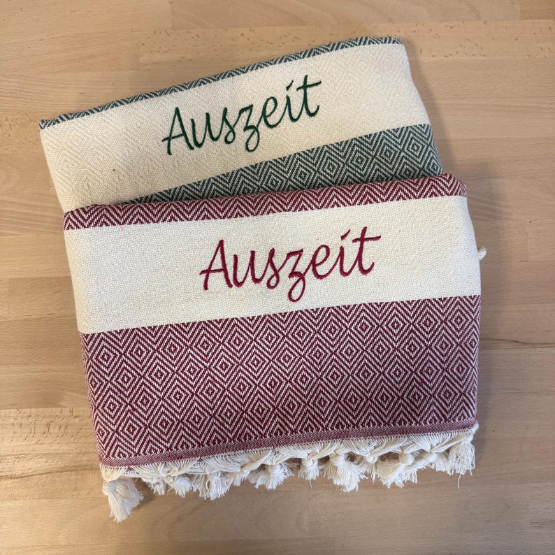 May include: Two folded Turkish towels with the word "Auszeit" embroidered in green and burgundy. The towels have a cream-colored center stripe and diamond pattern borders in green and burgundy. The towels have a fringe trim.