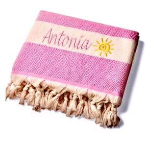 Könnte beinhalten: Ein gefaltetes rosa und cremefarbenes Textil mit einem rautenförmigen geometrischen Muster. Der Stoff enthält ein besticktes cremefarbenes Band mit dem Namen Antonia in rosa Schreibschrift und einem gelben Sonnensymbol, abgeschlossen mit geknoteten Fransen.