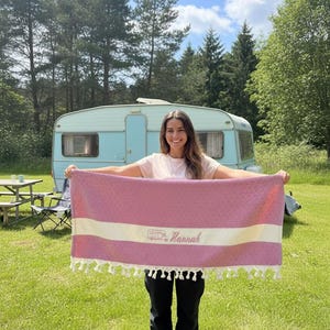 May include: A pink and white striped towel with a fringe, personalised with the name "Hannah" and a camper graphic. The towel is held in front of a light blue caravan in a grassy outdoor setting.