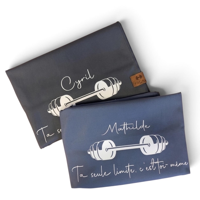 May include: Two folded rectangular towels in dark gray and blue-gray. Each features a white barbell graphic, a name in script font, and the text "Ta seule limite, c'est toi-m&ecirc;me". A small brown tag with a barbell icon and "FitGifts" is attached.