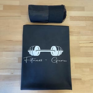 May include: A black rectangular mat featuring a white graphic of a barbell and the text Fitness - Guru in a script font, accompanied by a matching black cylindrical carrying case with a mesh section at one end for storage and transport.