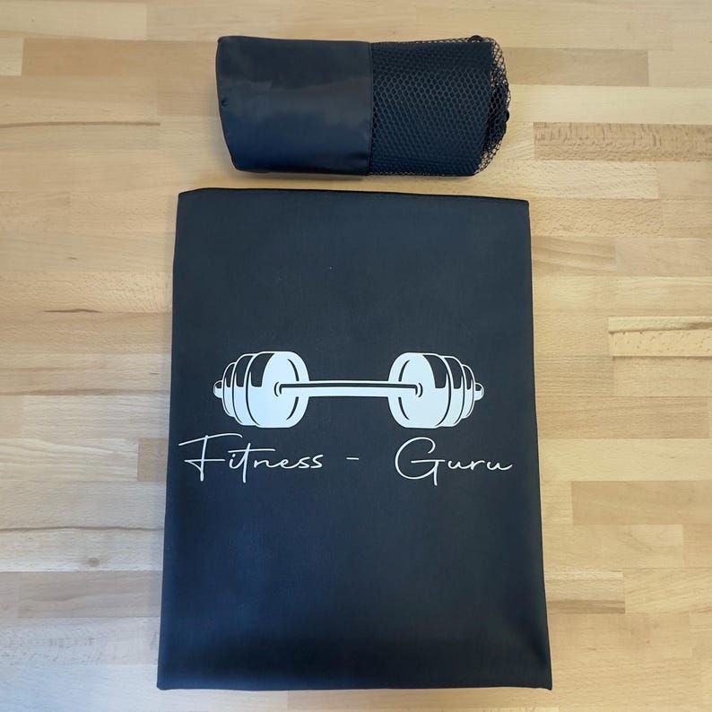 May include: A black rectangular mat featuring a white graphic of a barbell and the text Fitness - Guru in a script font, accompanied by a matching black cylindrical carrying case with a mesh section at one end for storage and transport.