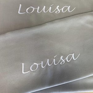 Può includere: Due pannelli di tessuto grigio con il nome "Louisa" ricamato in bianco su ogni pannello.