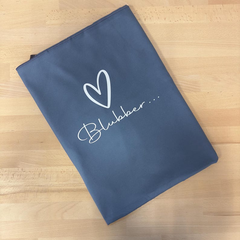May include: A rectangular slate blue zippered pouch featuring a white hand-drawn heart outline centered above the word Blubber... written in a white cursive script font. The pouch has a smooth, matte finish and a simple, clean design.