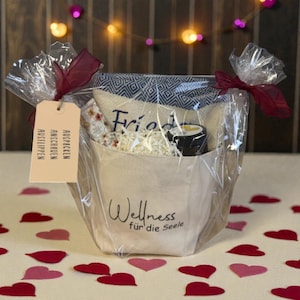 Könnte beinhalten: Ein Geschenkekorb, verpackt in durchsichtigem Zellophan und mit bordeauxfarbenen Bändern gebunden. Der Korb enthält eine kleine Canvas-Tasche mit dem Aufdruck "Wellness für die Seele", ein gemustertes Handtuch und andere kleine Artikel. Ein Anhänger mit deutschem Text hängt am Band.