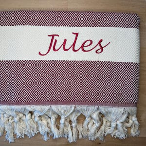 K&ouml;nnte beinhalten: Ein gefaltetes rechteckiges Textil mit einem geometrischen Rautenmuster in Dunkelrot und Cremewei&szlig;. Ein breites, cremewei&szlig;es Querband in der Mitte zeigt den in roter Schreibschrift gestickten Namen Jules. Die Unterkante ist mit geknoteten Fransen versehen.