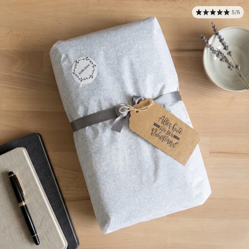 May include: A gift wrapped in light gray paper, tied with a gray ribbon and a brown tag that reads "Alles Gute f&uuml;r den Ruhestand". A small sticker with the words "with love" is on the wrapping. A pen and notebooks are in the background.