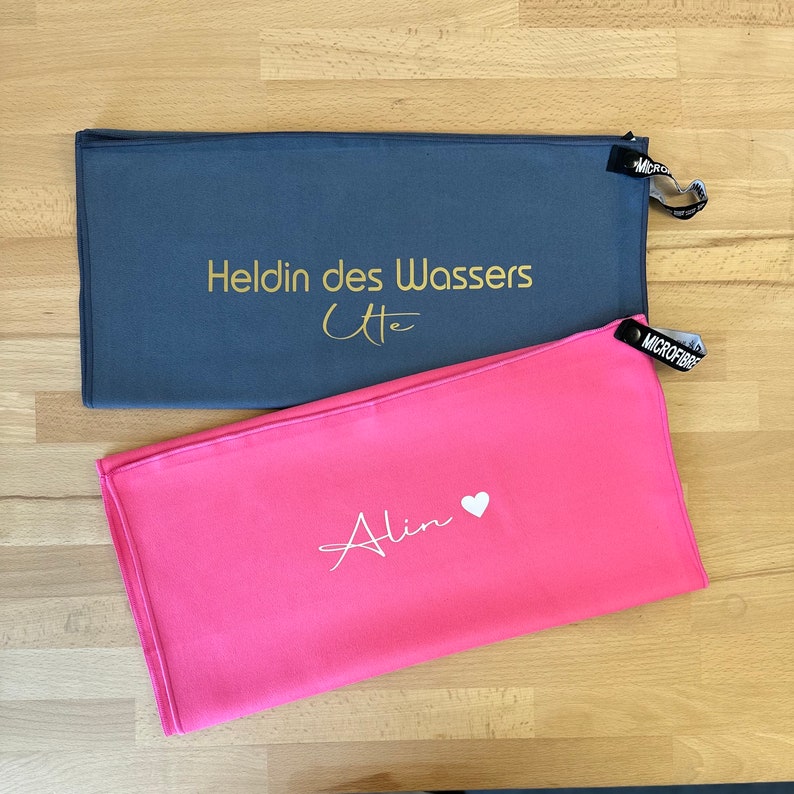 Pu&ograve; includere: Due asciugamani in microfibra, uno grigio con testo dorato che dice "Heldin des Wassers Ute" e uno rosa con testo bianco che dice "Alin" con un simbolo di cuore.