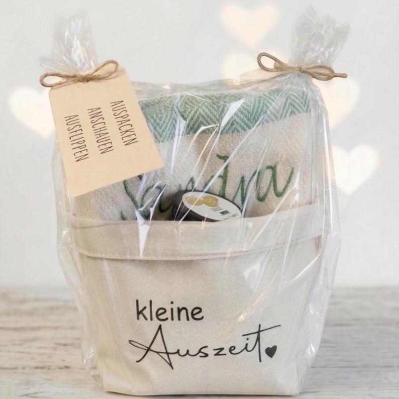 May include: A gift basket wrapped in clear cellophane, tied with twine. The basket contains a cream-colored towel with green accents and the name "Ava" embroidered in green. A tag reads "Auspacken, Anschauen, Ausflippen". The basket has the words "kleine Auszeit" printed on it.