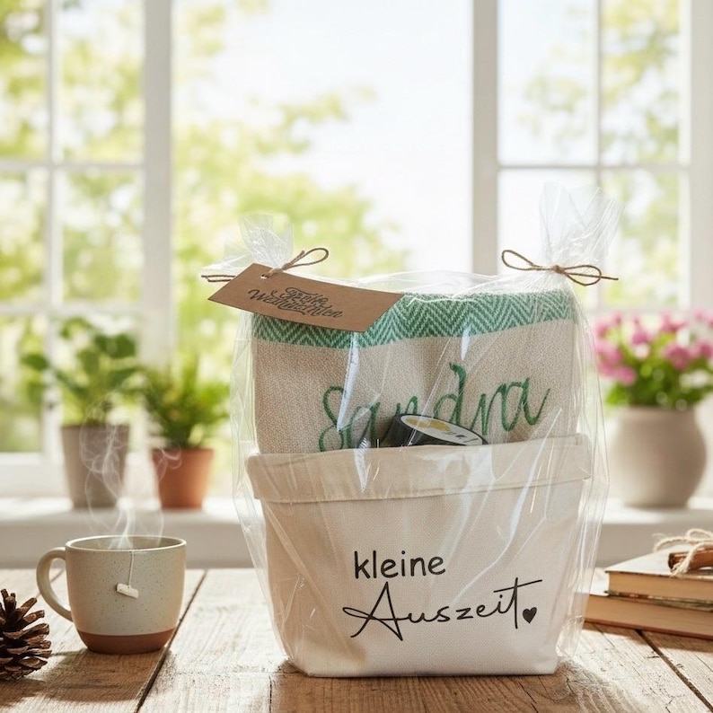 May include: A gift basket with a white canvas container and a green and white towel. The towel is embroidered with the name "Sandra". The container has the text "kleine Auszeit" and a heart. A cup of tea and a pinecone are in the foreground.
