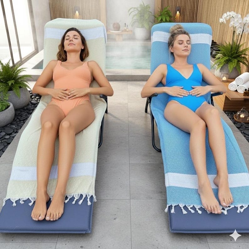 K&ouml;nnte beinhalten: Zwei Liegest&uuml;hle mit schwarzem Metallrahmen und marineblauen Polstern, jeweils bedeckt mit einem gro&szlig;en rechteckigen Handtuch. Ein Handtuch ist hellbeige mit wei&szlig;en Streifen und Fransen, das andere ist leuchtend blau mit wei&szlig;en Streifen und Fransen.