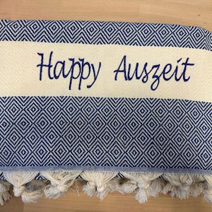 K&ouml;nnte beinhalten: Ein gefaltetes rechteckiges Textil mit einem dunkelblau-wei&szlig;en Rautenmuster. Ein breiter, cremefarbener horizontaler Streifen verl&auml;uft durch die Mitte und ist mit dem dunkelblauen Schriftzug Happy Auszeit bestickt. Die Unterkante hat cremefarbene Quasten.