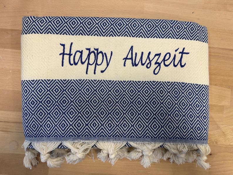 K&ouml;nnte beinhalten: Ein gefaltetes rechteckiges Textil mit einem dunkelblau-wei&szlig;en Rautenmuster. Ein breiter, cremefarbener horizontaler Streifen verl&auml;uft durch die Mitte und ist mit dem dunkelblauen Schriftzug Happy Auszeit bestickt. Die Unterkante hat cremefarbene Quasten.