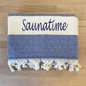 K&ouml;nnte beinhalten: Ein gefaltetes rechteckiges Textil mit einem blau-wei&szlig;en Rautenmuster. Der obere Bereich besteht aus einem cremefarbenen Band, auf dem das Wort Saunatime in dunkelblauer Schreibschrift gestickt ist. Die Unterkante ist mit wei&szlig;en Fransen versehen.