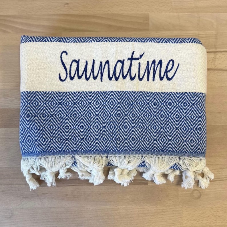 K&ouml;nnte beinhalten: Ein gefaltetes rechteckiges Textil mit einem blau-wei&szlig;en Rautenmuster. Der obere Bereich besteht aus einem cremefarbenen Band, auf dem das Wort Saunatime in dunkelblauer Schreibschrift gestickt ist. Die Unterkante ist mit wei&szlig;en Fransen versehen.