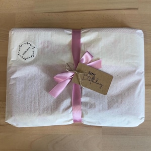 K&ouml;nnte beinhalten: Ein rechteckiges Paket, eingewickelt in wei&szlig;es Seidenpapier und mit einem hellrosa Satinband zu einer Schleife gebunden. Ein runder Aufkleber zeigt den Schriftzug "with love" in einem Kranz, und ein braunes Etikett tr&auml;gt den Text "Happy Birthday".