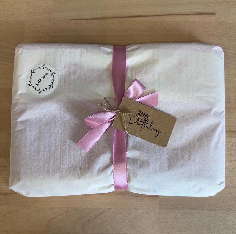 K&ouml;nnte beinhalten: Ein rechteckiges Paket, eingewickelt in wei&szlig;es Seidenpapier und mit einem hellrosa Satinband zu einer Schleife gebunden. Ein runder Aufkleber zeigt den Schriftzug "with love" in einem Kranz, und ein braunes Etikett tr&auml;gt den Text "Happy Birthday".