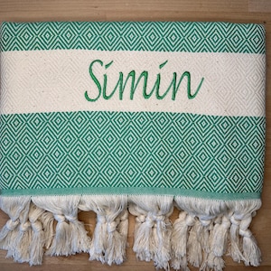 May include: A green and white patterned towel with the name "S&iacute;min" embroidered in green. The towel features a diamond pattern and cream-colored tassels along the bottom edge. The towel is folded and lies on a wooden surface.