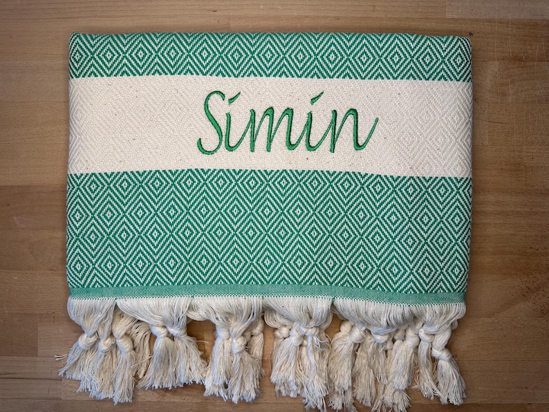 May include: A green and white patterned towel with the name "S&iacute;min" embroidered in green. The towel features a diamond pattern and cream-colored tassels along the bottom edge. The towel is folded and lies on a wooden surface.