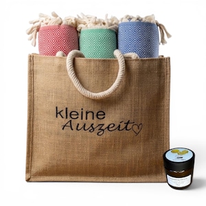 Pode incluir: Uma bolsa de juta bege com alças de corda, apresentando o texto preto "kleine Auszeit" e um coração. Dentro, três toalhas enroladas em vermelho, verde e azul com padrões de diamante e franjas. Um pequeno frasco de vidro âmbar está ao lado.