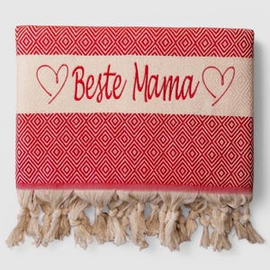 May include: A red and cream-colored Turkish towel with a diamond pattern. The towel features the embroidered text "Beste Mama" in red, framed by heart shapes. Cream-colored fringe adorns the bottom edge.