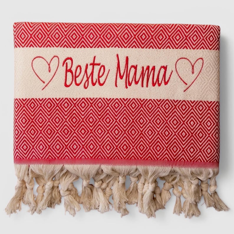 May include: A red and cream-colored Turkish towel with a diamond pattern. The towel features the embroidered text "Beste Mama" in red, framed by heart shapes. Cream-colored fringe adorns the bottom edge.