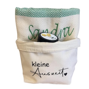 May include: A white canvas bag with the text "kleine Auszeit" and a heart. Inside is a folded towel with green chevron trim and the name "Sandra" embroidered in green. A black container with a white label is inside the bag.