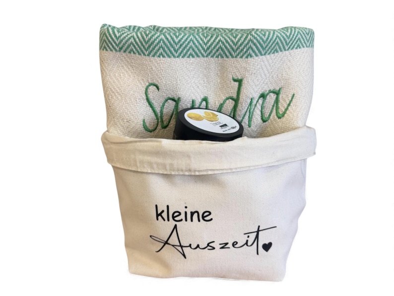 May include: A white canvas bag with the text "kleine Auszeit" and a heart. Inside is a folded towel with green chevron trim and the name "Sandra" embroidered in green. A black container with a white label is inside the bag.