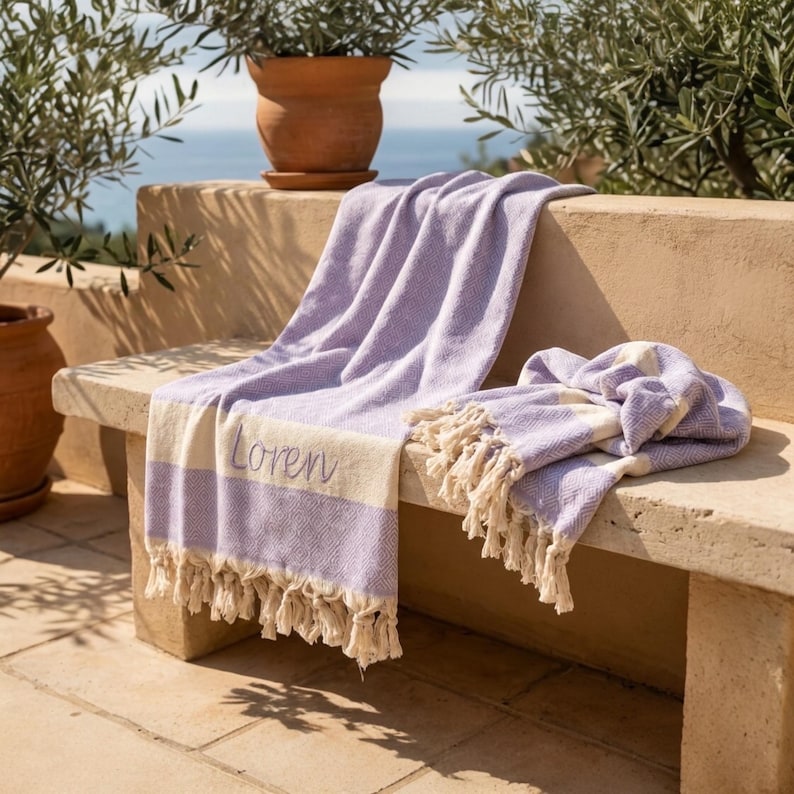 May include: A lavender and cream-colored beach towel with the word "Lovern" embroidered on it. The towel has a fringed edge and is draped over a stone bench. A second towel is folded on the bench. The background includes potted plants and a view of the ocean.