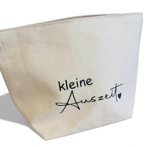May include: Cream-colored canvas tote bag with the words "kleine Auszeit" in black script. The bag has a simple, tapered design and a small heart symbol at the end of the text.