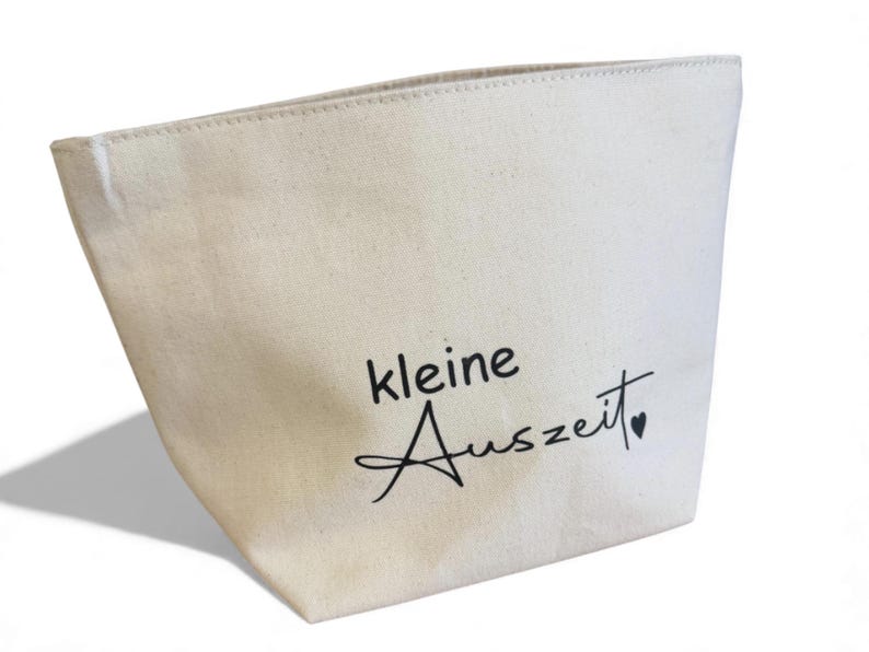 May include: Cream-colored canvas tote bag with the words "kleine Auszeit" in black script. The bag has a simple, tapered design and a small heart symbol at the end of the text.