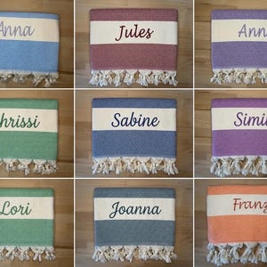 K&ouml;nnte beinhalten: Eine Sammlung von neun rechteckigen Webhandt&uuml;chern mit einem zentralen wei&szlig;en horizontalen Streifen und eingestickten Namen wie Anna, Jules, Anni, Chrissi, Sabine, Simin, Lori, Joanna und Franzi. Jedes Handtuch hat ein strukturiertes Muster und wei&szlig;e Quasten.
