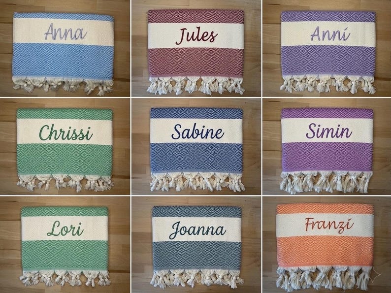 K&ouml;nnte beinhalten: Eine Sammlung von neun rechteckigen Webhandt&uuml;chern mit einem zentralen wei&szlig;en horizontalen Streifen und eingestickten Namen wie Anna, Jules, Anni, Chrissi, Sabine, Simin, Lori, Joanna und Franzi. Jedes Handtuch hat ein strukturiertes Muster und wei&szlig;e Quasten.