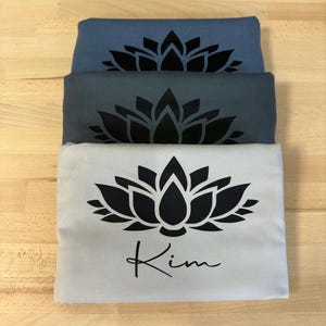 May include: Three folded t-shirts stacked vertically. The top shirt is light blue, the middle is dark gray, and the bottom is light gray. Each shirt features a black lotus flower graphic. The bottom shirt also includes the name Kim in black script.