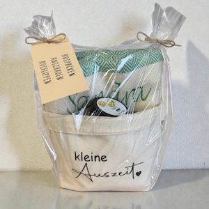 May include: A gift basket wrapped in clear cellophane. Inside is a small canvas bucket with the words "kleine Auszeit" in black script. The basket contains a folded green and white patterned blanket, a small black item, and a tag with German text.