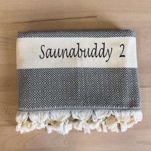 K&ouml;nnte beinhalten: Ein gefaltetes schwarz-wei&szlig;es Handtuch mit einem sich wiederholenden Rautenmuster und einem einfarbigen Streifen, der den gestickten Text "Saunabuddy 2" in einer schwarzen Schreibschrift zeigt. Der untere Rand ist mit geknoteten Fransen versehen.
