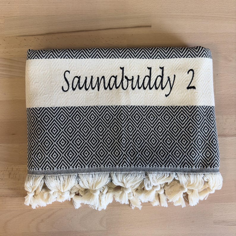 K&ouml;nnte beinhalten: Ein gefaltetes schwarz-wei&szlig;es Handtuch mit einem sich wiederholenden Rautenmuster und einem einfarbigen Streifen, der den gestickten Text "Saunabuddy 2" in einer schwarzen Schreibschrift zeigt. Der untere Rand ist mit geknoteten Fransen versehen.