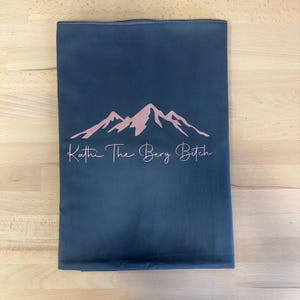 May include: A rectangular dark gray fabric pouch featuring a rose gold mountain range graphic centered above the text Kathi The Berg Bitch in a cursive script font. The item is flat and displays a clean, smooth surface with a zipper closure at the top.