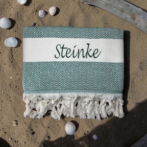 K&ouml;nnte beinhalten: Ein gefaltetes rechteckiges Textil mit einem dunkelgr&uuml;nen und wei&szlig;en Rautenmuster und einem durchgehenden wei&szlig;en horizontalen Streifen in der Mitte. Das Wort Steinke ist in dunkelgr&uuml;ner Schreibschrift auf den wei&szlig;en Streifen gestickt. Fransen am Rand.