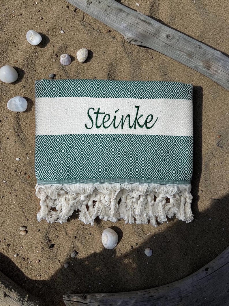 K&ouml;nnte beinhalten: Ein gefaltetes rechteckiges Textil mit einem dunkelgr&uuml;nen und wei&szlig;en Rautenmuster und einem durchgehenden wei&szlig;en horizontalen Streifen in der Mitte. Das Wort Steinke ist in dunkelgr&uuml;ner Schreibschrift auf den wei&szlig;en Streifen gestickt. Fransen am Rand.