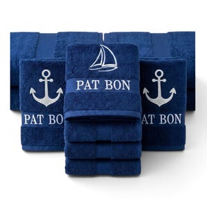 Könnte beinhalten: Ein Set dunkelblauer Handtücher mit weißen, gestickten nautischen Motiven. Die Handtücher zeigen ein Segelboot-Symbol über dem Text PAT BON, während andere ein Anker-Symbol über dem Text PAT BON aufweisen. Der Stoff hat eine weiche Textur.