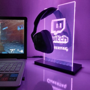Könnte beinhalten: Ein violettes LED-Licht mit einem schwarzen Headset und dem Twitch-Logo. Das Licht steht auf einem schwarzen Sockel und befindet sich auf einer weißen Oberfläche.