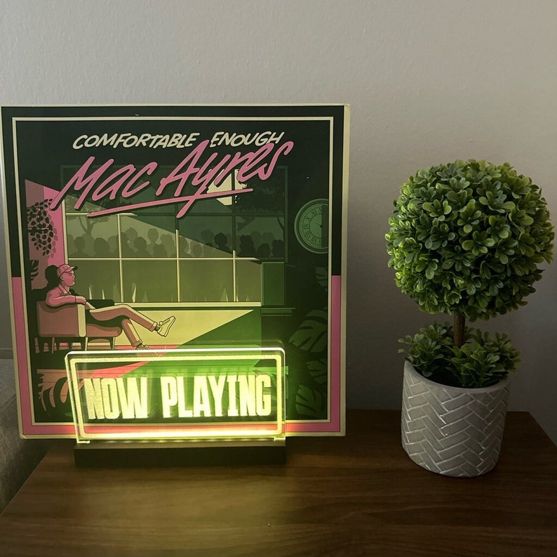 K&ouml;nnte beinhalten: Eine gr&uuml;ne und schwarze Schallplatte mit dem Titel "Comfortable Enough Mac Ayres" und einem gr&uuml;nen Neon-Schild mit der Aufschrift "NOW PLAYING".