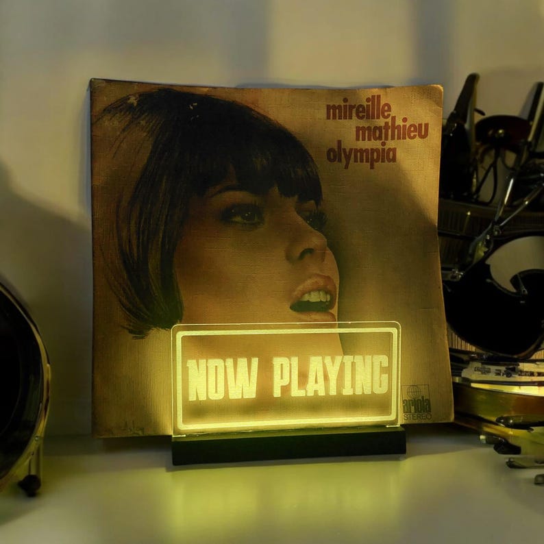 K&ouml;nnte beinhalten: Ein gelbes Neon-Schild mit den Worten "NOW PLAYING" in wei&szlig;en Buchstaben. Das Schild steht auf einem schwarzen Sockel und befindet sich vor einem Vintage-Schallplattenalbum.