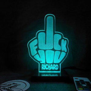 Puede incluir: Un dedo medio luminoso azul turquesa con la palabra "F**K YOU" y el nombre "RICHARD" debajo. La luz está sobre una base negra y tiene un mando a distancia.