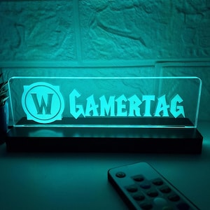 以下が含まれることがあります： 黒い土台に付けられたターコイズブルーのアクリル製の看板。看板には白い「W」のロゴと、スタイリッシュなフォントで「GAMERTAG」という文字が書かれている。