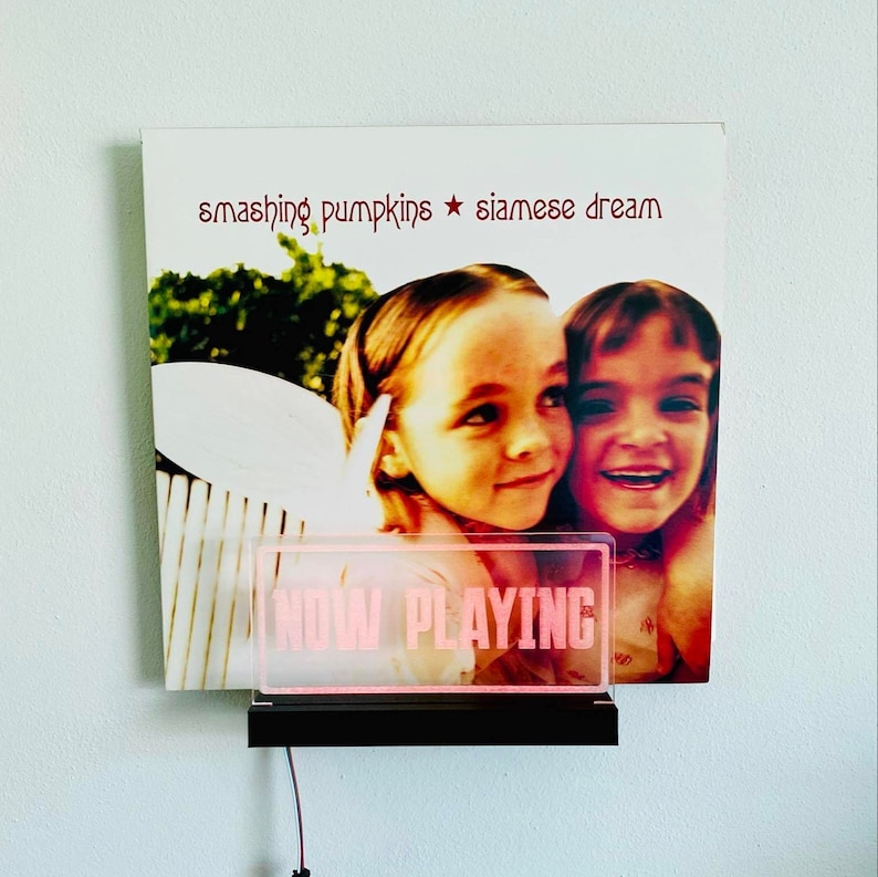 K&ouml;nnte beinhalten: Ein Vinyl-Plattencover mit einem Foto von zwei jungen M&auml;dchen, die l&auml;cheln. Das Album tr&auml;gt den Titel "Smashing Pumpkins Siamese Dream". Vor dem Albumcover befindet sich ein pinkfarbenes Neon-Schild mit der Aufschrift "NOW PLAYING".