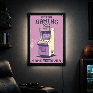Puede incluir: Un letrero enmarcado de un club de juegos retro con un fondo púrpura. El letrero presenta una ilustración crema y púrpura de una máquina recreativa vintage. El texto "RETRO GAMING Club" y "GAME is not OVER" se muestran en blanco.